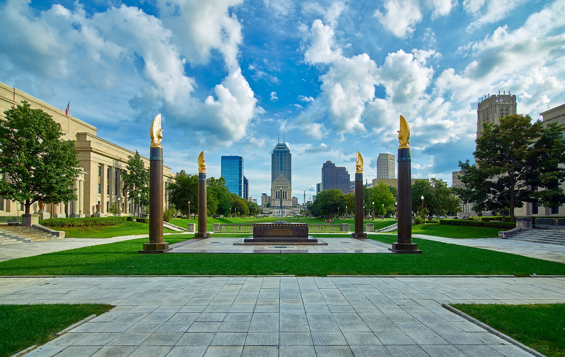 Image of the American Legion Mall in Indianapolis, with the skyline in the background. There is a bright blue sky, and bright green grass, in the center is a memorial, surrounded by 4 columns.